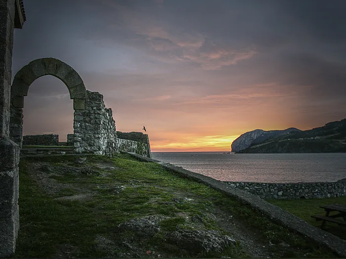 Un arco de piedra sobre el prado en Mundaka, con el mar y el cielo de atardecer al fondo.